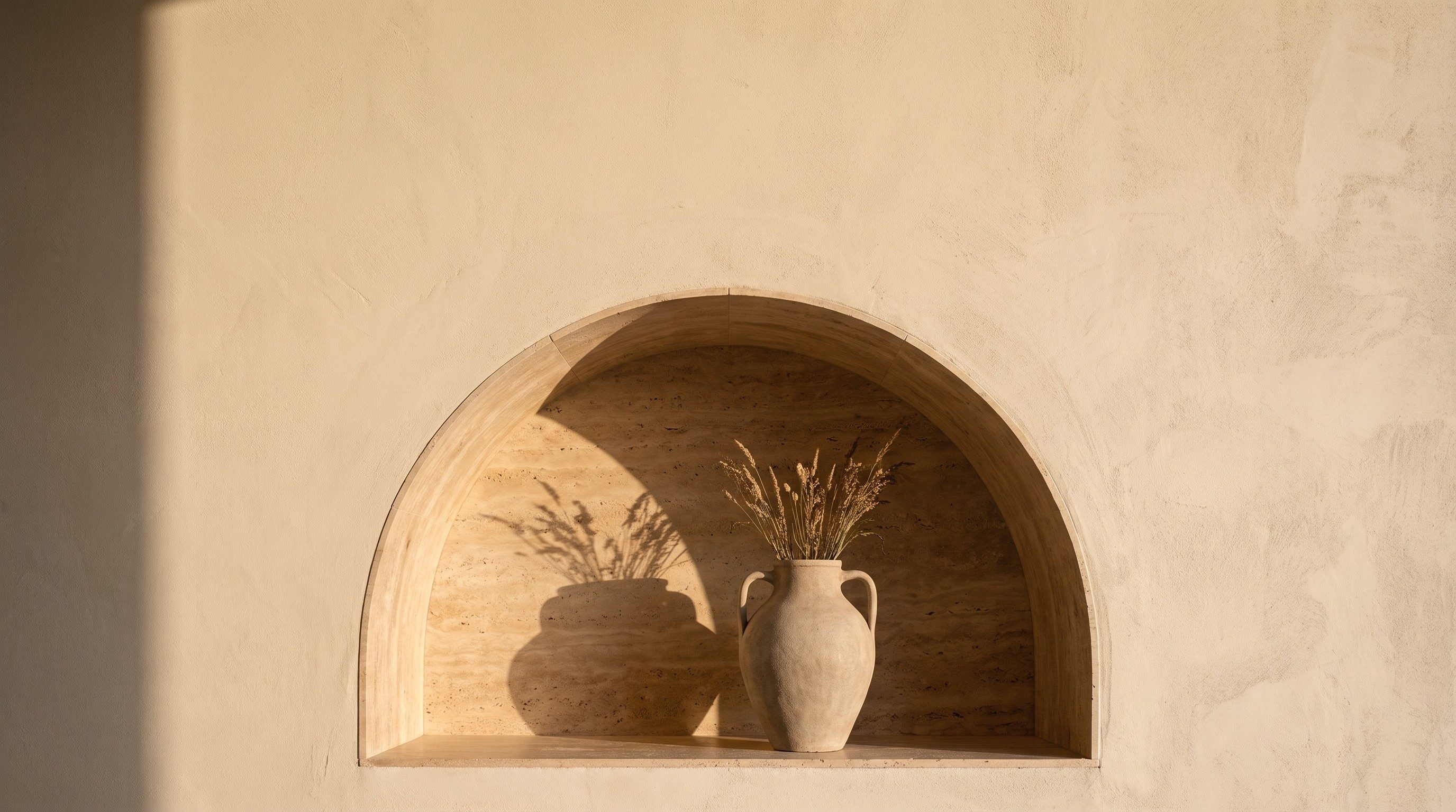 An arched niche in a warm lime-plaster wall, a stone amphora of dried grasses inside it, low sun laying its shadow across the travertine.