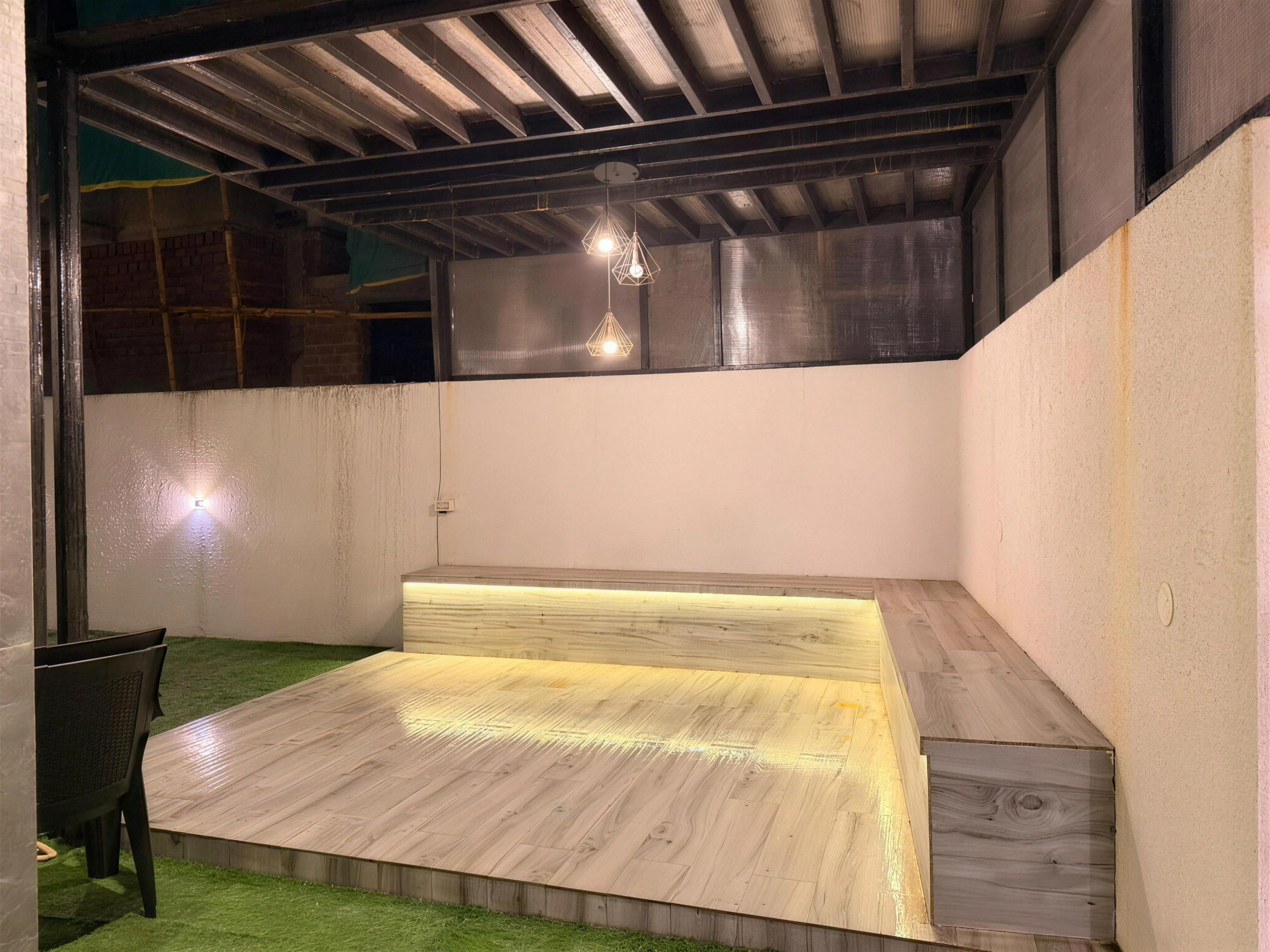 A rooftop deck at night under a cold cast: a timber platform with a lit bench, a pale wall and a stray chair at the left edge of the frame.