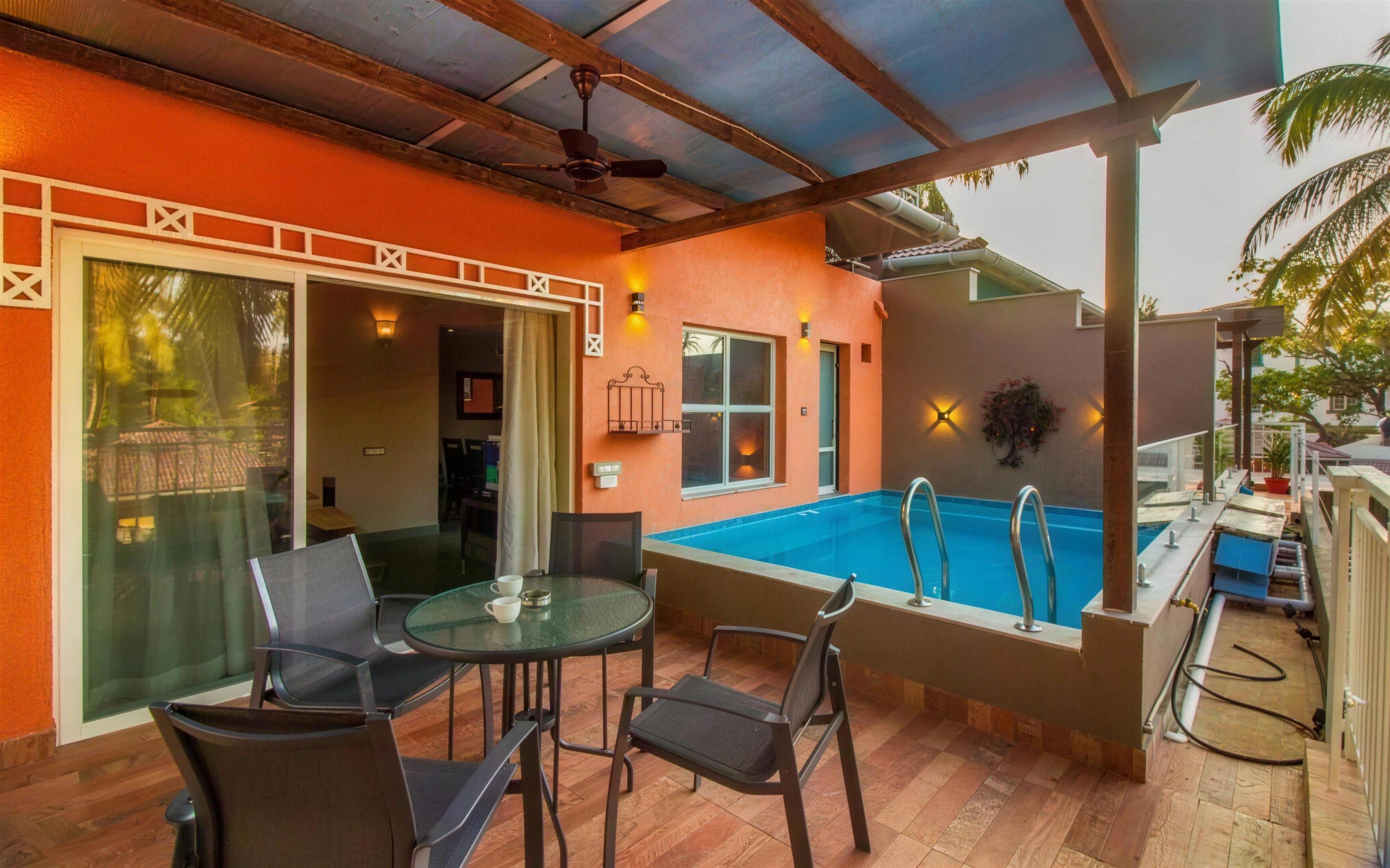 A covered terrace in hard daylight: an orange wall, a bright blue plunge pool, a glass table with four chairs and a hose left coiled on the deck.