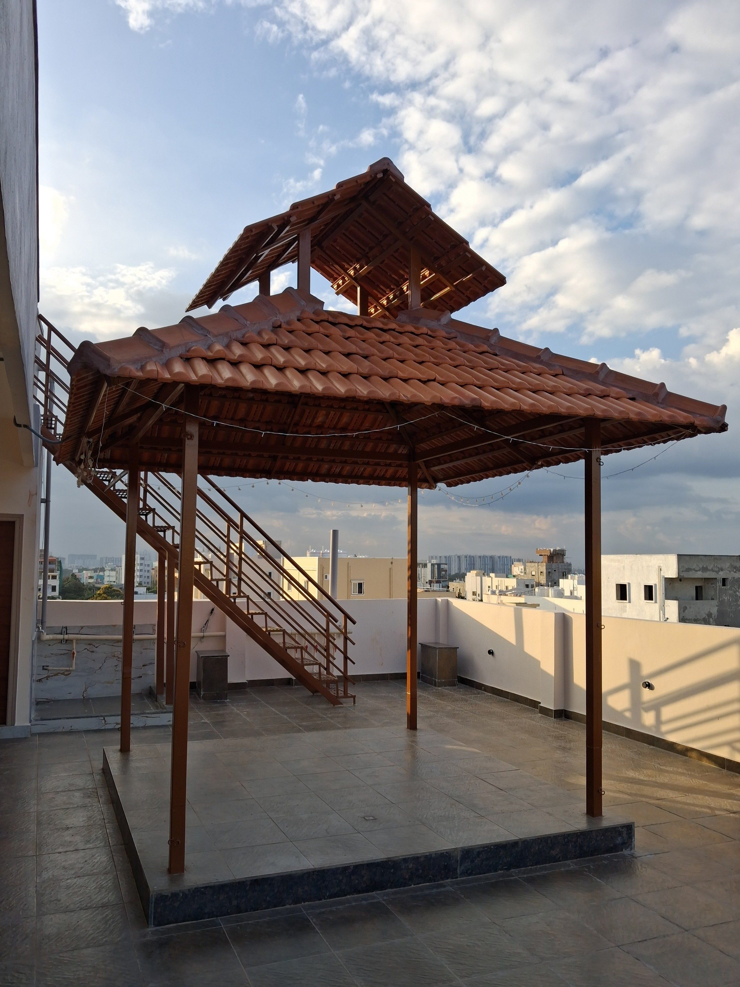 A rooftop terrace photographed in portrait and into the light: a tiled gazebo with a raised cupola and string lights on four slim posts, a steel staircase rising behind it on the left, and a cream parapet with neighbouring buildings beyond.